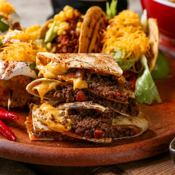A wooden board piled with cheesy quesadillas, tacos, and red chili peppers from El Castillo Mexicano.