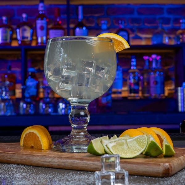 A large margarita glass with ice and an orange slice, surrounded by lime and orange wedges.